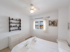 A bedroom with a bed, a dresser, a shelf with decorative items, a ceiling fan, and a window with curtains. - Apartment  by Interhome (Nerja)