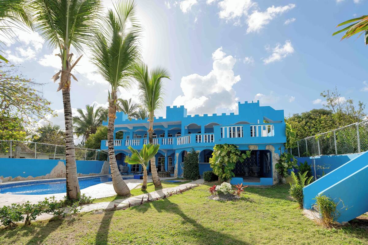 Un edificio azul con piscina, rodeado de palmeras y follaje.