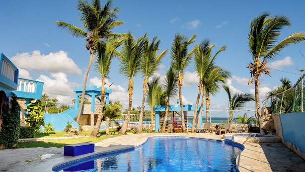 A swimming pool with palm trees, a blue building, and a view of the ocean. - Hotel Pirata Boca de Yuma (Boca de Yuma)