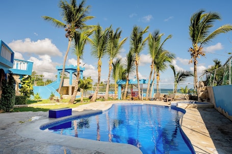 A swimming pool with palm trees, a blue building, and a view of the ocean.