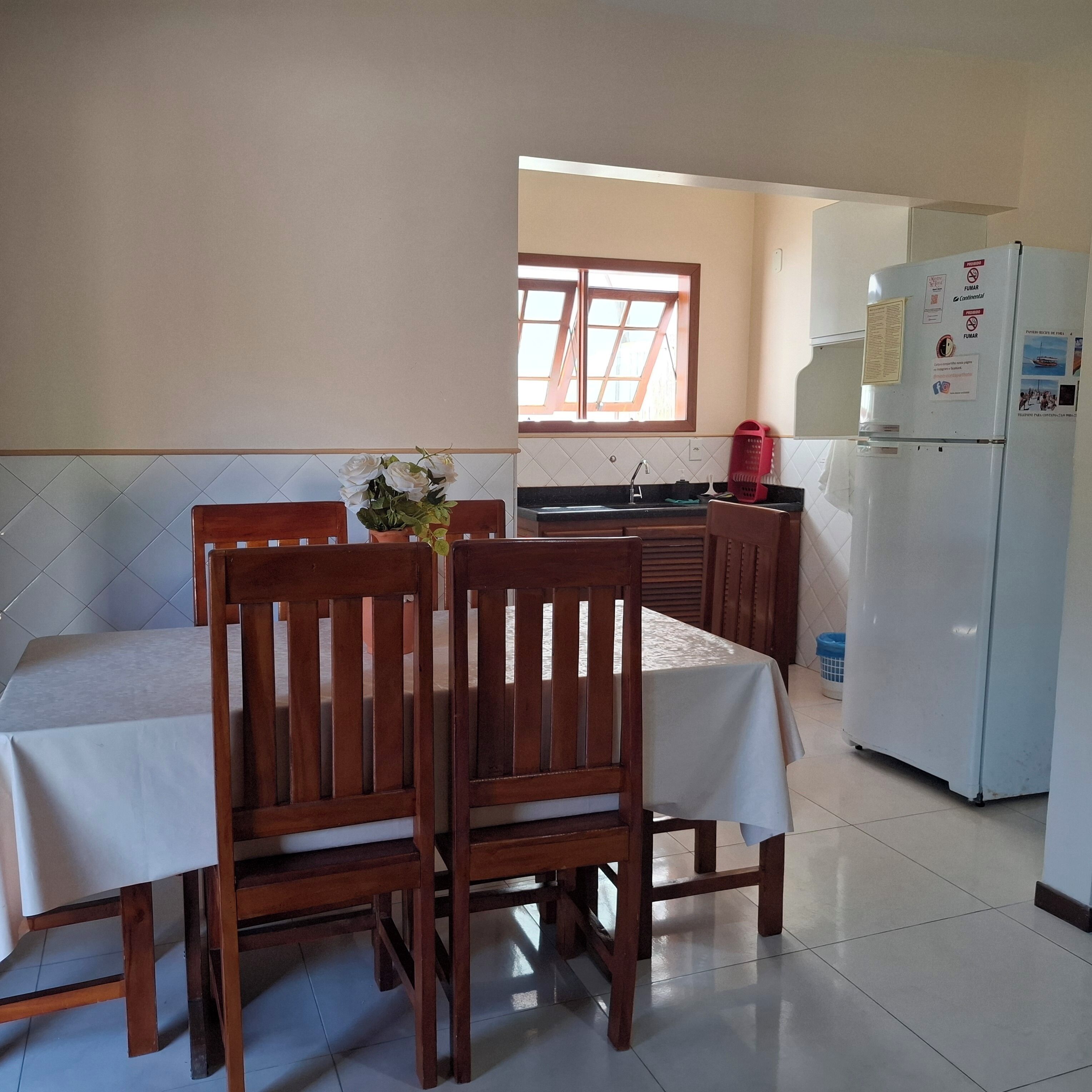 Un comedor con mesa y sillas, una ventana con persianas, una zona de cocina con refrigerador y un piso de baldosas.