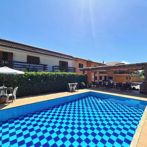 A swimming pool with a checkered pattern, outdoor seating, and a building with a tiled roof. - Mestre Lord aparthotel by La Vem Ferias  (Porto Seguro)