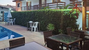 A poolside area with a table and chairs, a hanging light fixture, and a building with a balcony. - Mestre Lord aparthotel by La Vem Ferias  (Porto Seguro)