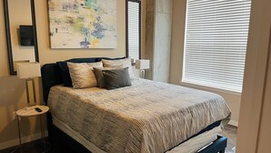 A neatly made bed with a patterned bedspread, a bedside lamp, a wall-mounted TV, and a large abstract painting. - Quiet Enjoyment in Houston Medical Center (Houston)