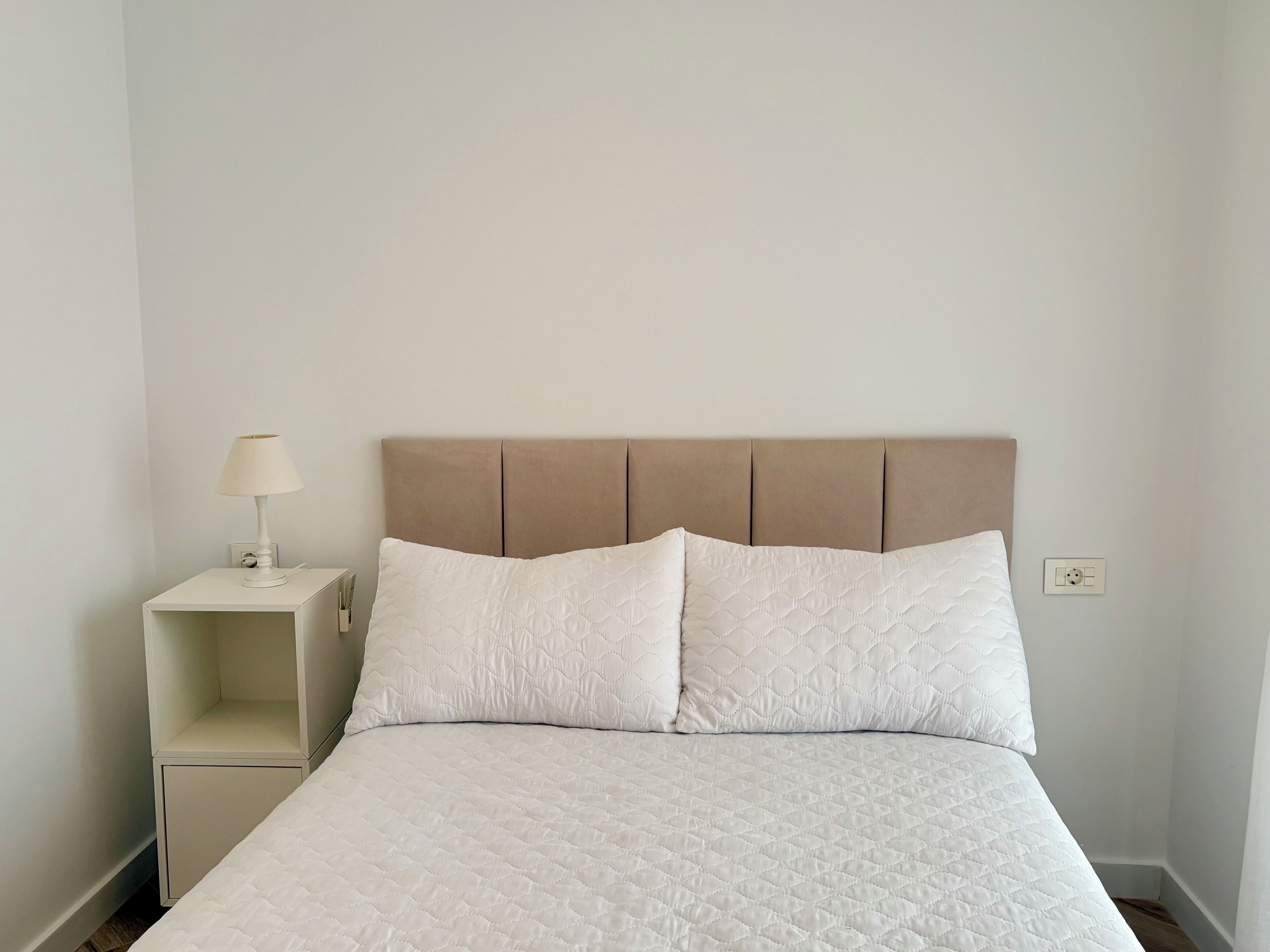 Un lit avec un matelas blanc matelassé, une tête de lit avec des panneaux verticaux, une table de chevet avec une lampe et une prise électrique sur le mur.