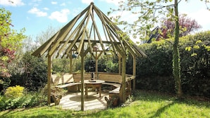 Een houten tuinpaviljoen met een bank en tafel, omringd door begroeiing en een stenen muur.