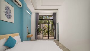 A bedroom with a bed, a nightstand, a wall-mounted air conditioner, a window with curtains, and a view of greenery outside. - Cam On Luxury Pool 4BR Villa by the Beach (Da Nang)
