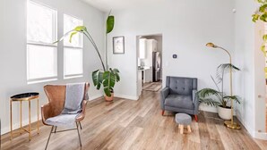 Un salon spacieux avec un canapé gris, un fauteuil marron, une table basse ronde et un grand lampadaire. De grandes fenêtres, une plante en pot et une vue sur une autre pièce complètent l’ensemble.