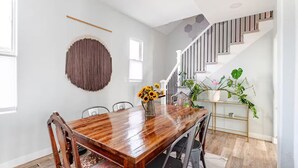 Un coin repas avec une table en bois, des chaises, un élément décoratif suspendu au mur, un escalier et des plantes en pot.