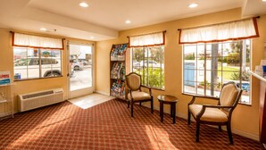 Eine Hotelhalle mit einem gemusterten Teppich, einer Rezeption, einem Sessel, einem kleinen Tisch und großen Fenstern mit Vorhängen.