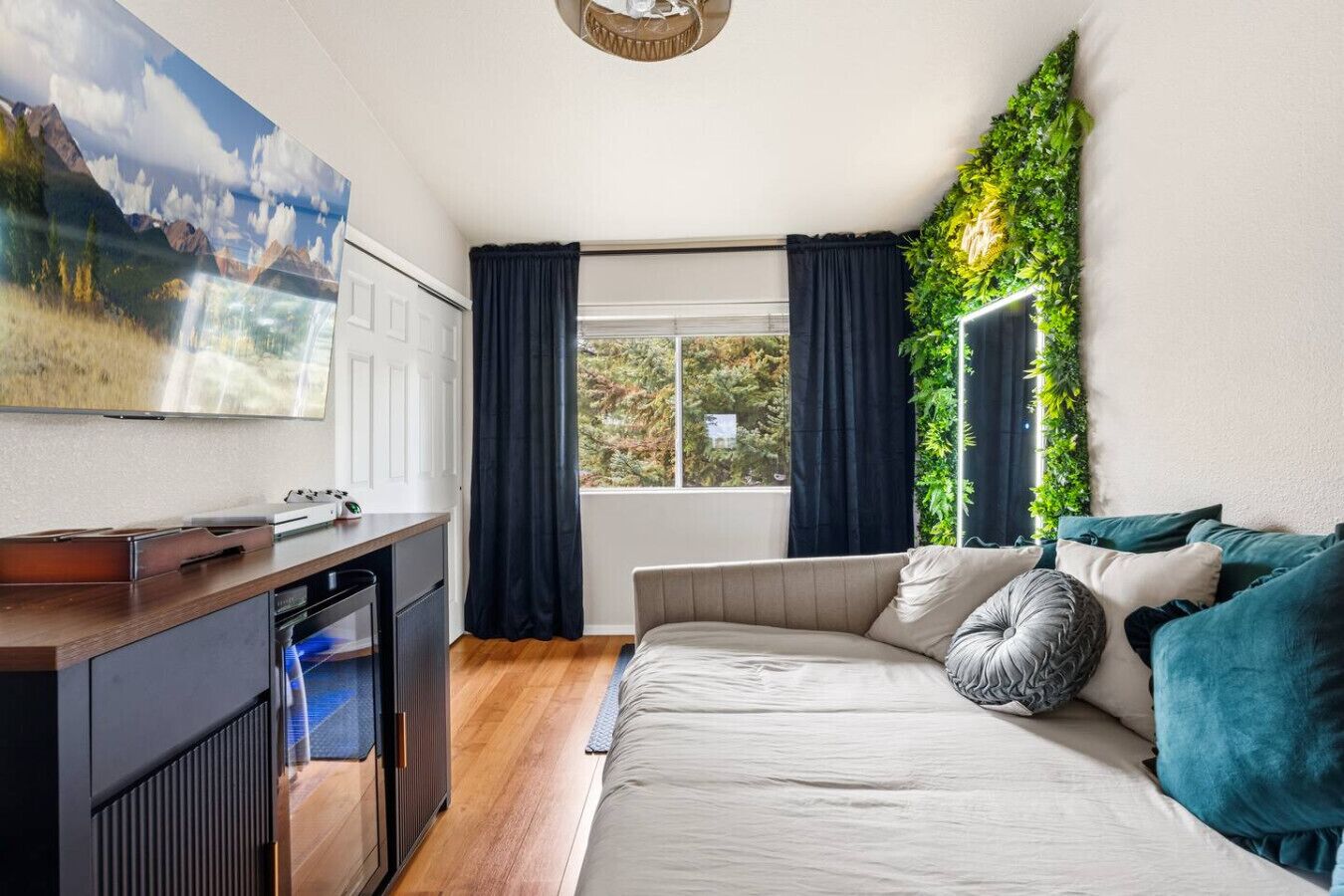 A room with a bed, a desk with a computer, a window with curtains, and a wall-mounted plant.