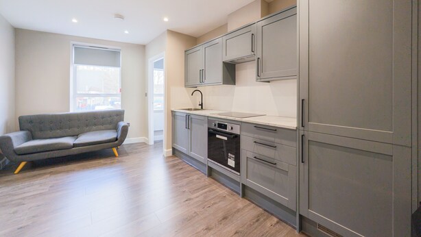 A modern kitchen with grey cabinets, a built-in oven, and a grey sofa in a spacious living area with wooden flooring. - Harbour View Apartments Portsmouth (Portsmouth)