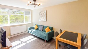 A living room with a sofa, a table, a TV, and a window with a view of trees. - Harbour View Apartments Portsmouth (Portsmouth)