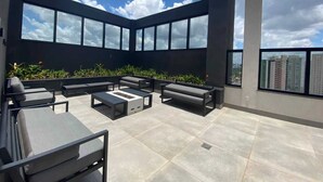A rooftop terrace with a modern design, featuring a seating area with sofas and a table, surrounded by large windows offering a view of the cityscape. - Cozy Apartment - Prime Building (Campo Grande)