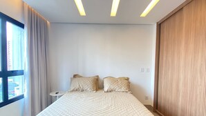 A modern bedroom with a large bed, bedside tables, a window with curtains, and a wooden panel wall. - Cozy Apartment - Prime Building (Campo Grande)