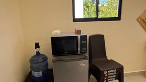 A compact kitchenette with a refrigerator, microwave, and water dispenser on a small table. A chair is positioned next to the table, and a window provides a view of greenery outside.