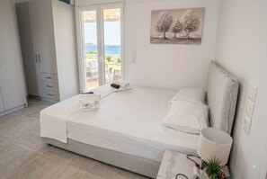 A bedroom with a bed, a bedside table with a lamp, a painting on the wall, and a view of the outdoors through a sliding door. - Sadova Messinia Pearl Cozy Seafront Tiny House (Kitries)
