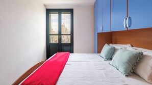 Una camera da letto con un letto, una coperta rossa, cuscini verde chiaro e una testiera in legno, oltre a una finestra che offre vista su edifici.