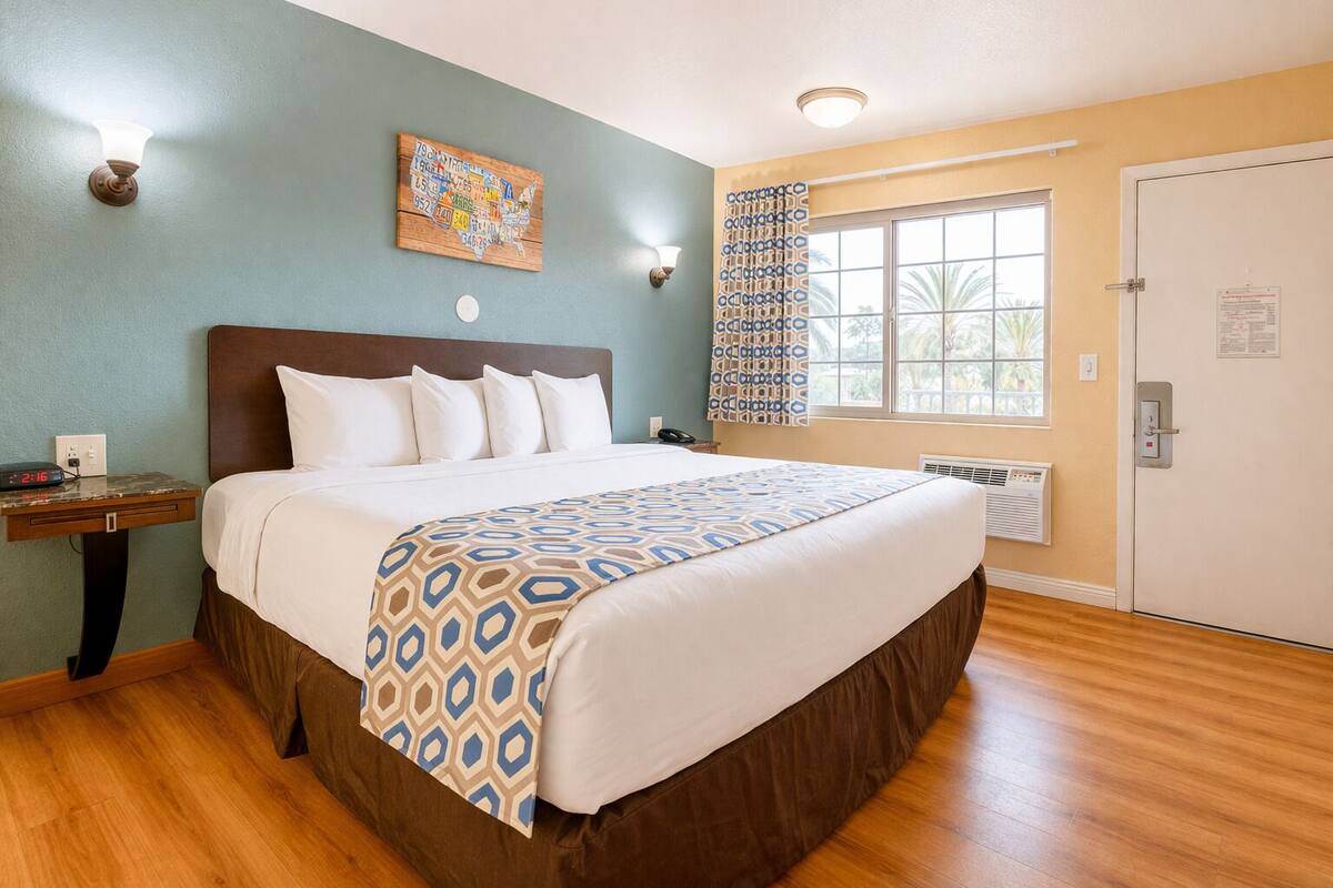 A hotel room with a bed, a nightstand, a wall clock, a window with a view, and a door.
