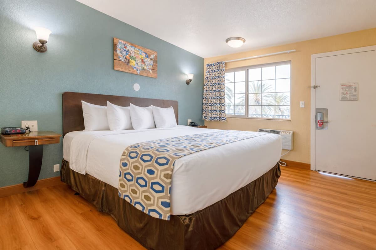 A hotel room with a bed, a nightstand, a wall clock, a window with a view, and a door.