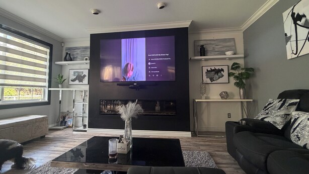 Ein Wohnzimmer mit Flachbildfernseher, einer schwarzen Couch, einem Couchtisch und einem Kamin.