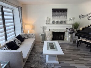 Ein Wohnzimmer mit einer Couch, einem Couchtisch, einem Kamin, einem Klavier und einem großen Fenster mit Jalousien.
