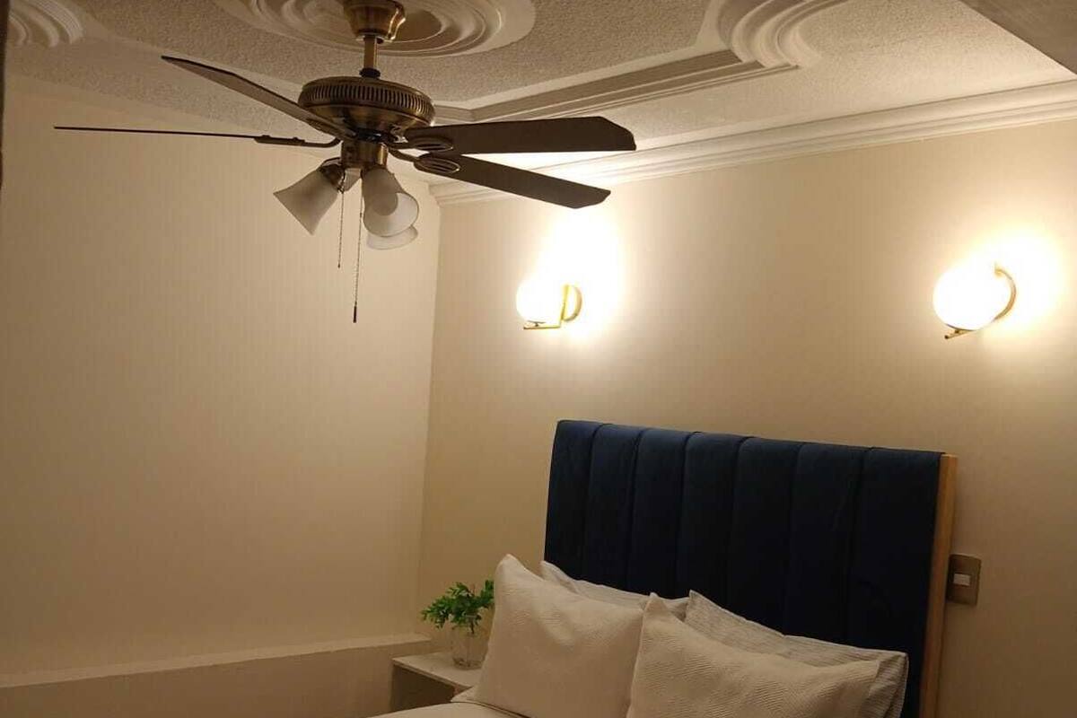 Habitación de hotel con una cama, ventilador de techo, apliques de pared y una mesita de noche con una planta.