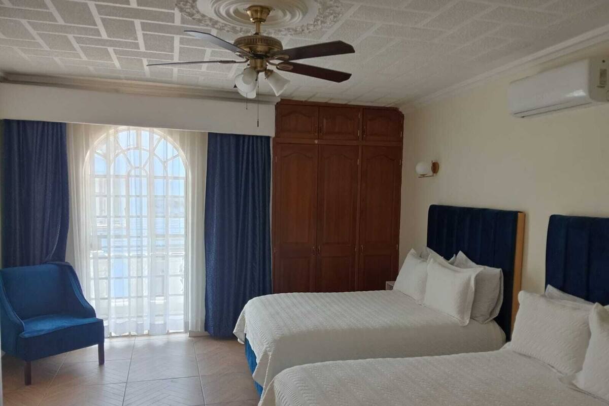 Habitación de hotel con dos camas, un ventilador de techo, un armario de madera, un sillón azul y una ventana con cortinas.