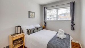 Quarto com cama, mesa de cabeceira com abajur, janela com cortinas e quadro na parede.