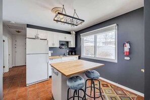 A kitchen with white cabinets, a wooden island, two bar stools, a hanging light fixture, and a fire extinguisher on the wall. - 80 min Banff, Minutes from Foothills Hospital (Calgary)