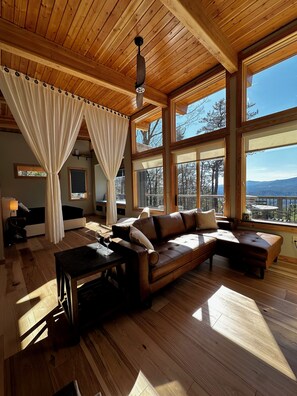 Una habitación acogedora con techo de madera, ventanas amplias, un sofá de cuero marrón y una mesa de comedor.