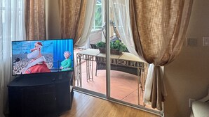 Un salon équipé d’un téléviseur, doté d’un plancher en bois, d’une porte-fenêtre coulissante et de rideaux.