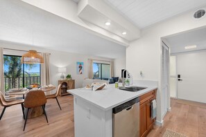 Een moderne keuken met een vaatwasser, een eethoek met een ronde tafel en stoelen, en uitzicht op palmbomen door grote ramen.