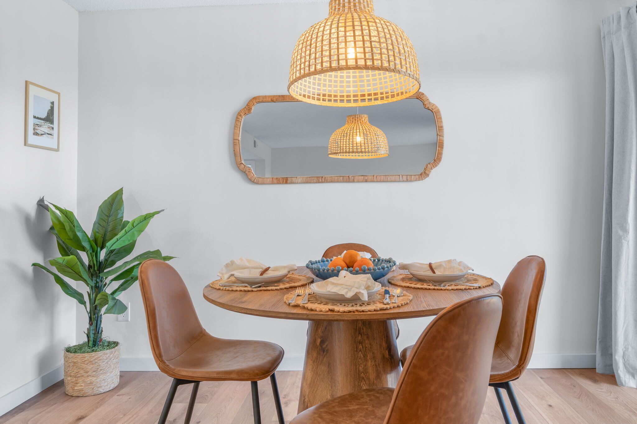 A dining area with a round wooden table, brown leather chairs, a woven pendant light, a mirror, and a potted plant.