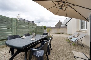 Een terras buiten met een tafel, stoelen en een grote parasol, met een grindpad.
