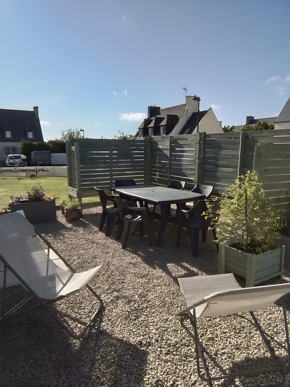 Eine Terrasse mit Tisch und Stühlen, umgeben von einem Holzzaun, Kies und Kübelpflanzen.