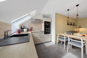A modern kitchen with a built-in oven, a coffee maker, and a dining area with a table and chairs. - Location 300 m from the Beach (Roscoff)
