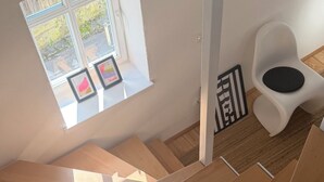 Ein modernes Interieur mit einer Holztreppe, einem kleinen Fenster mit Kunstwerk und einem weißen Stuhl mit schwarzer Sitzkissen.