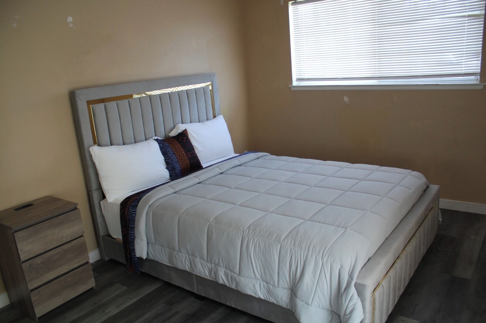 A neatly made bed with a headboard, a bedside table, and a window with blinds.