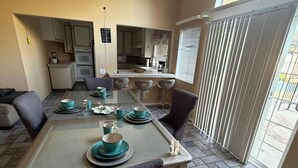 Un comedor con una mesa dispuesta para cuatro, una cocina al fondo y un amplio ventanal con persianas.