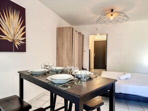 A dining area with a table set for four, a bed with a white duvet, and a door leading to another room.