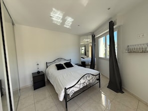 Ein Schlafzimmer mit Bett, Nachttisch, Spiegel, einem Fenster mit Vorhängen und einem Regal mit Kerzen.