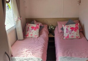 A compact room with two beds, each with pink bedding and butterfly-patterned pillows, a wooden headboard, and a small table with a vase of flowers. - D&T Caravan Hire Southview Holiday Park Skegness (Skegness)