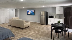 A modern living room with a sofa, a TV, a dining table with chairs, a kitchen area with cabinets, and a refrigerator. - Modern Open Studio - 3 Beds - 2 queens & 1–Full Sleeps 6- Near Local Attractions (Marksville)