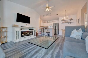 A modern living room with a glass coffee table, a grey sofa, a flat-screen TV, a ceiling fan, and a kitchen in the background. - Coastal 2BR Duplex – Near Beach, Parks, Dining & Free Trolley (Bradenton Beach)