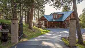 A log cabin with a metal roof, a stone gate with a 'WELCOME' sign, and a paved driveway leading to the entrance. - Beautiful In Bigfork - Newly built Studio Apmt. (Bigfork)