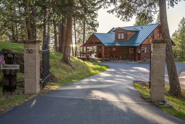 A log cabin with a metal roof, a stone gate with a 'WELCOME' sign, and a paved driveway leading to the entrance.