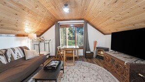 A cozy loft bedroom with a sloped ceiling, a large window, a fireplace, a sofa, a dining table, and a TV. - Beautiful In Bigfork - Newly built Studio Apmt. (Bigfork)