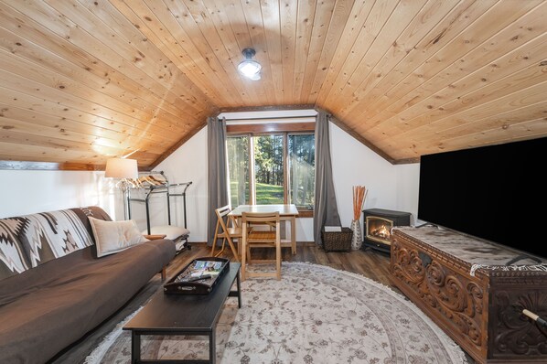 A cozy loft bedroom with a sloped ceiling, a large window, a fireplace, a sofa, a dining table, and a TV.