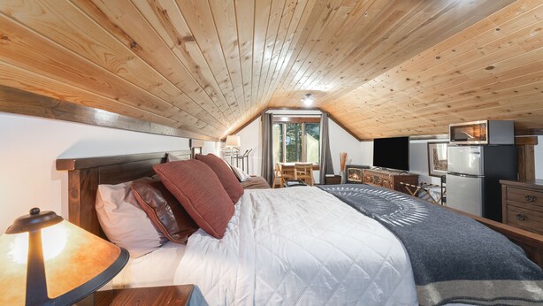 A bedroom with a wooden ceiling, a bed with pillows, a table with a lamp, a desk, a chair, a window, and a refrigerator. - Beautiful In Bigfork - Newly built Studio Apmt. (Bigfork)